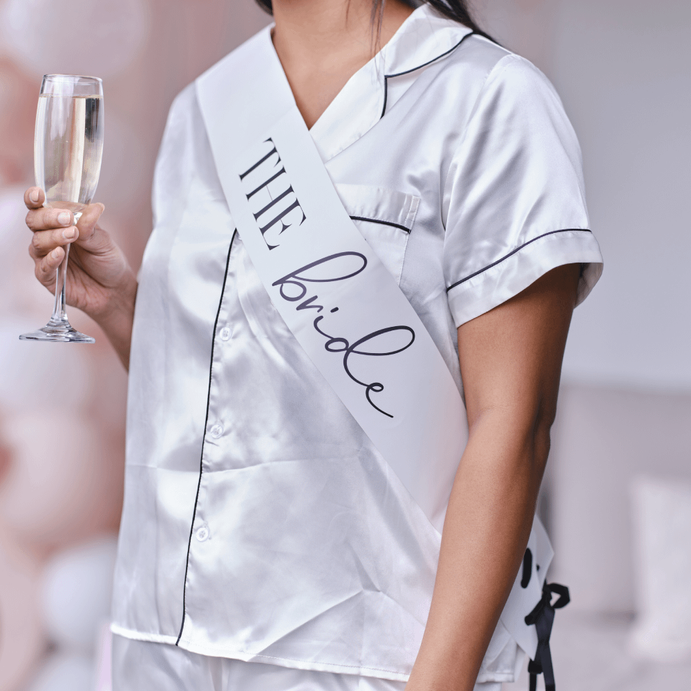 Vrouw in witte pyjama met een champagneglas en een sjerp met de tekst the bride