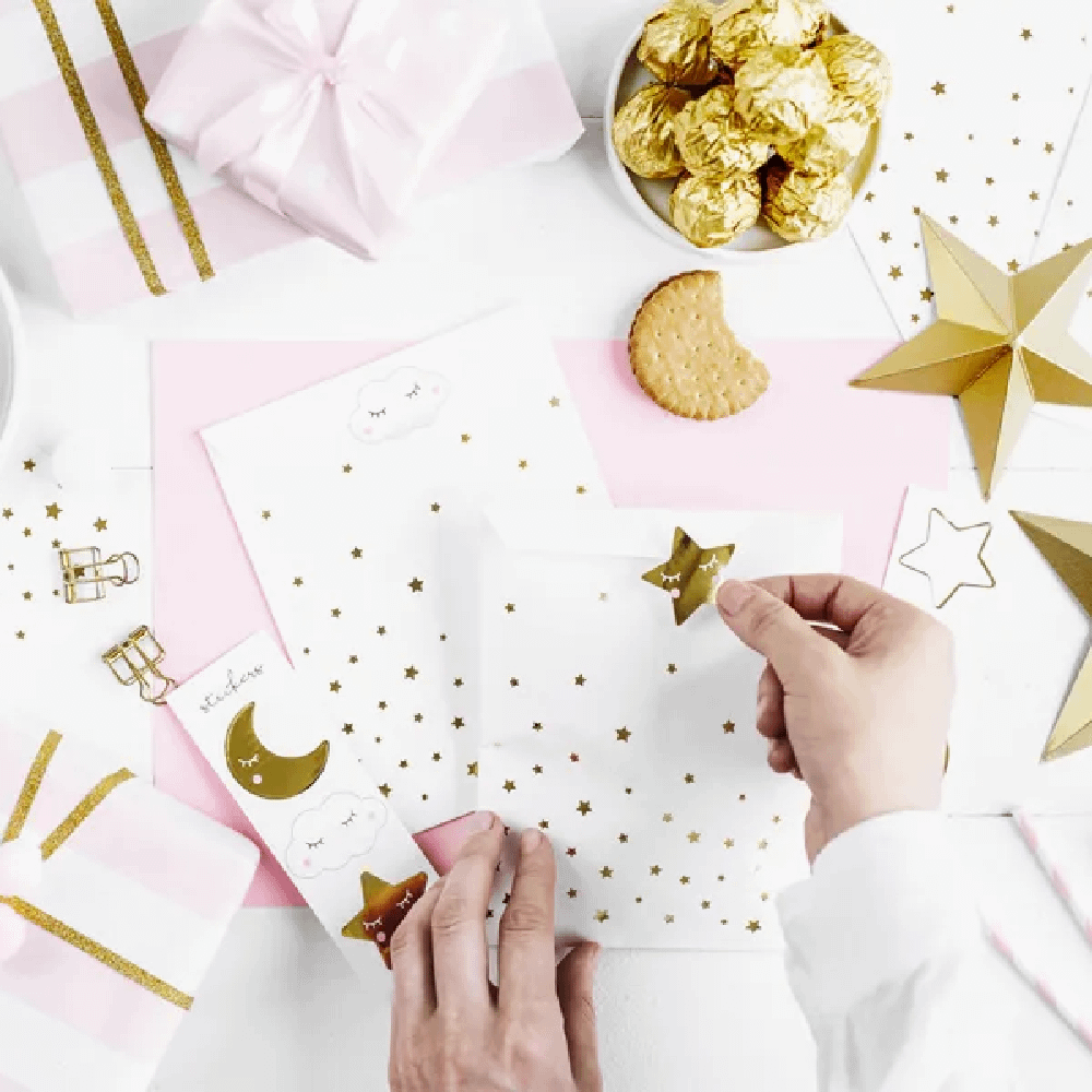 Witte zakjes met gouden sterren liggen op een lichtroze ondergrond met koekjes, bonbons in gouden verpakking en gouden paperclips