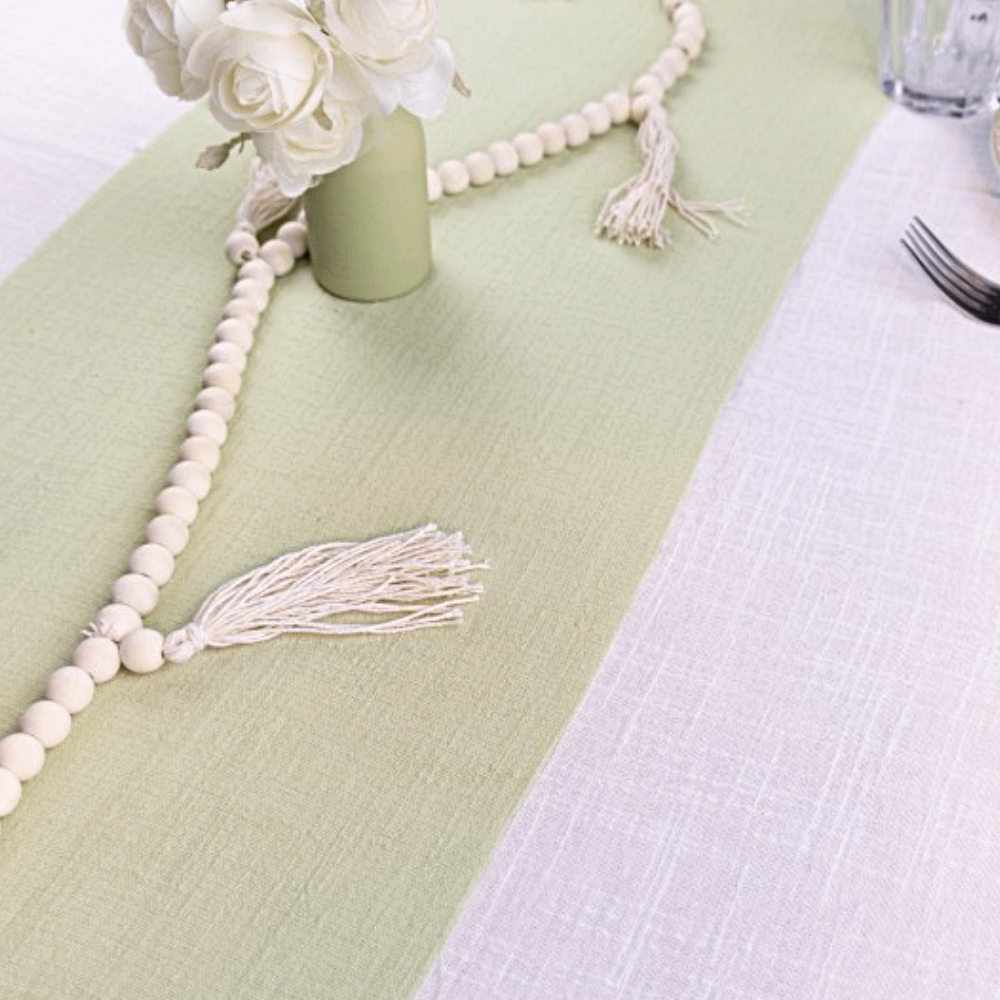 Saliegroene tafelloper ligt op een wit kleed en is versierd met een saliegroen vaasje en een kralenslinger