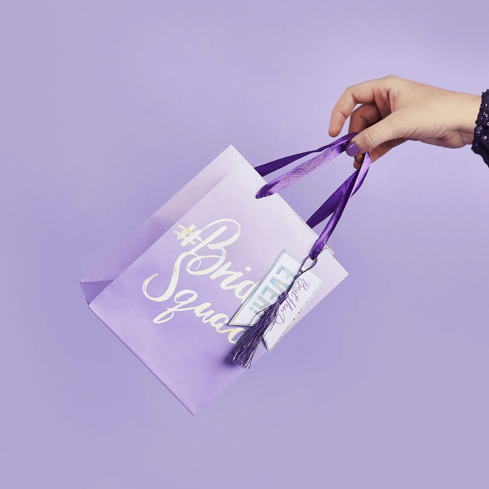 Hand houd een paars cadeautasje vast met iridescent tekst en een paarse tassel