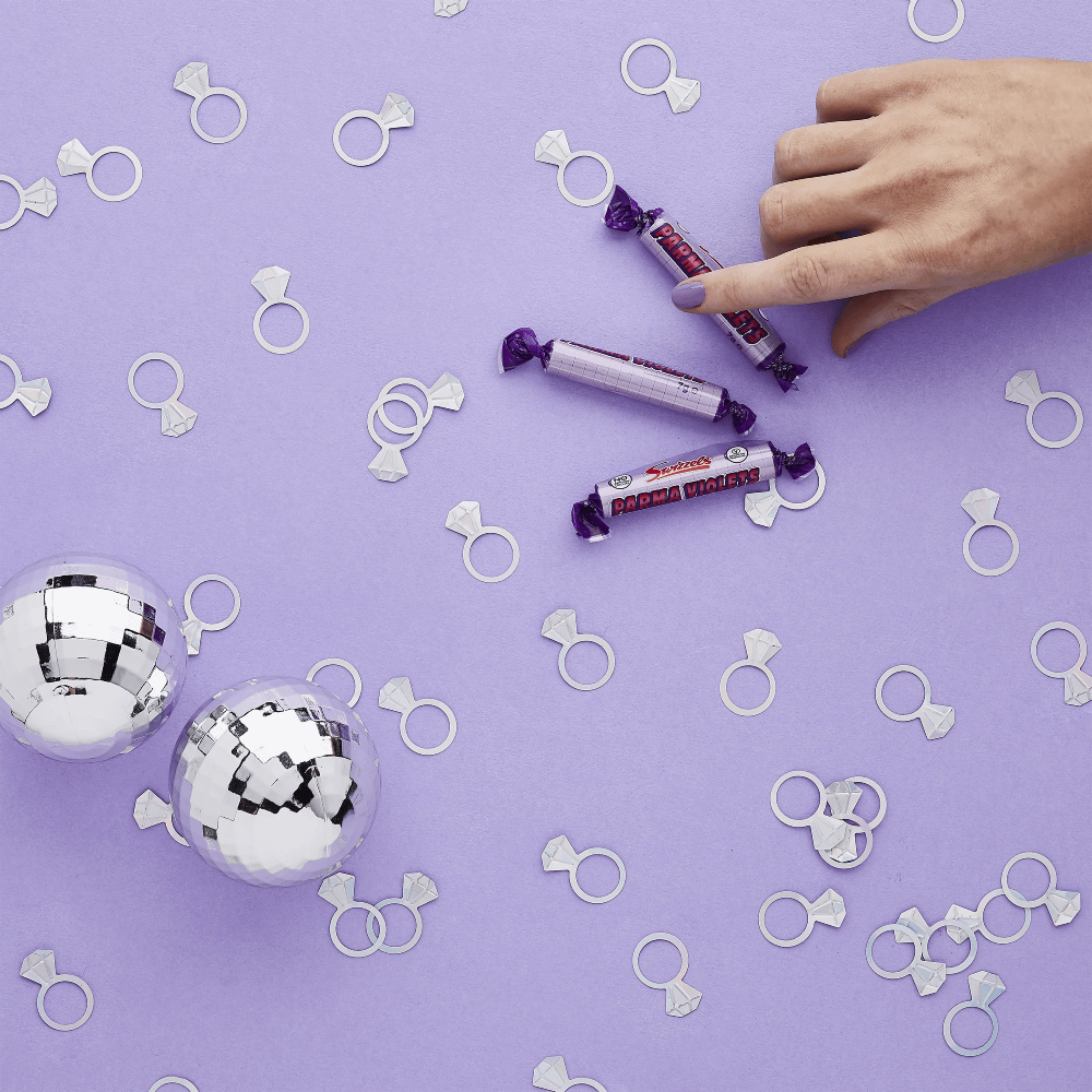 Iridescent confetti in de vorm van trouwringen ligt op een lila achtergrond naast paarse snoeprolletjes