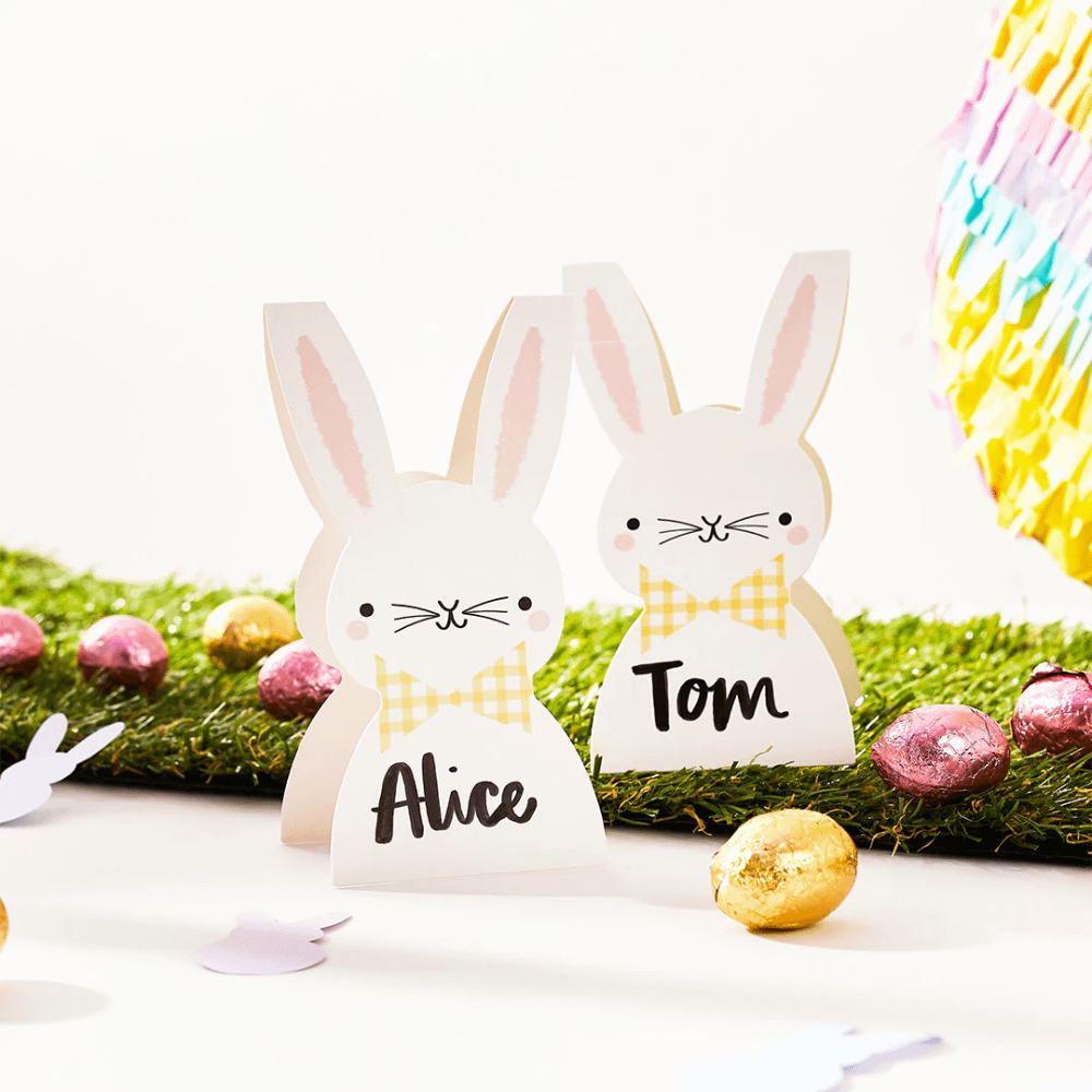 Tafelkaartjes in de vorm van konijntjes met een gele, geblokte strik om staan op gras versierd met gouden en roze paaseitjes
