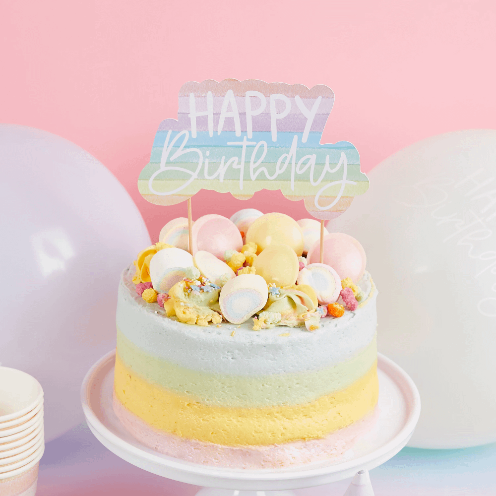 Taart topper met de tekst happy birthday in pasteltinten zit in een pastelkleurige regenboog taart op een wit plateau en staat voor ballonnen