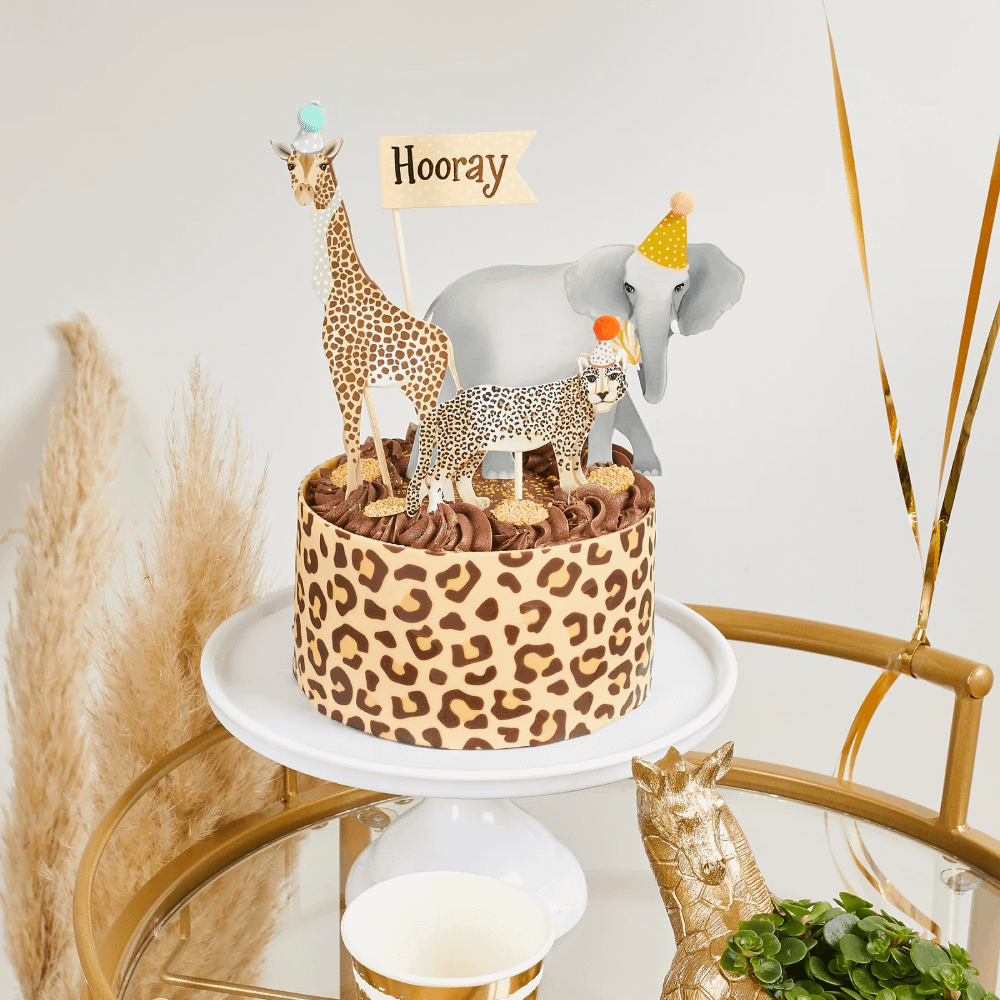 Caketoppers met een olifant, giraffe en tijger met feesthoedjes op staan op een chocoladetaart met tijgerprint op een wit plateau