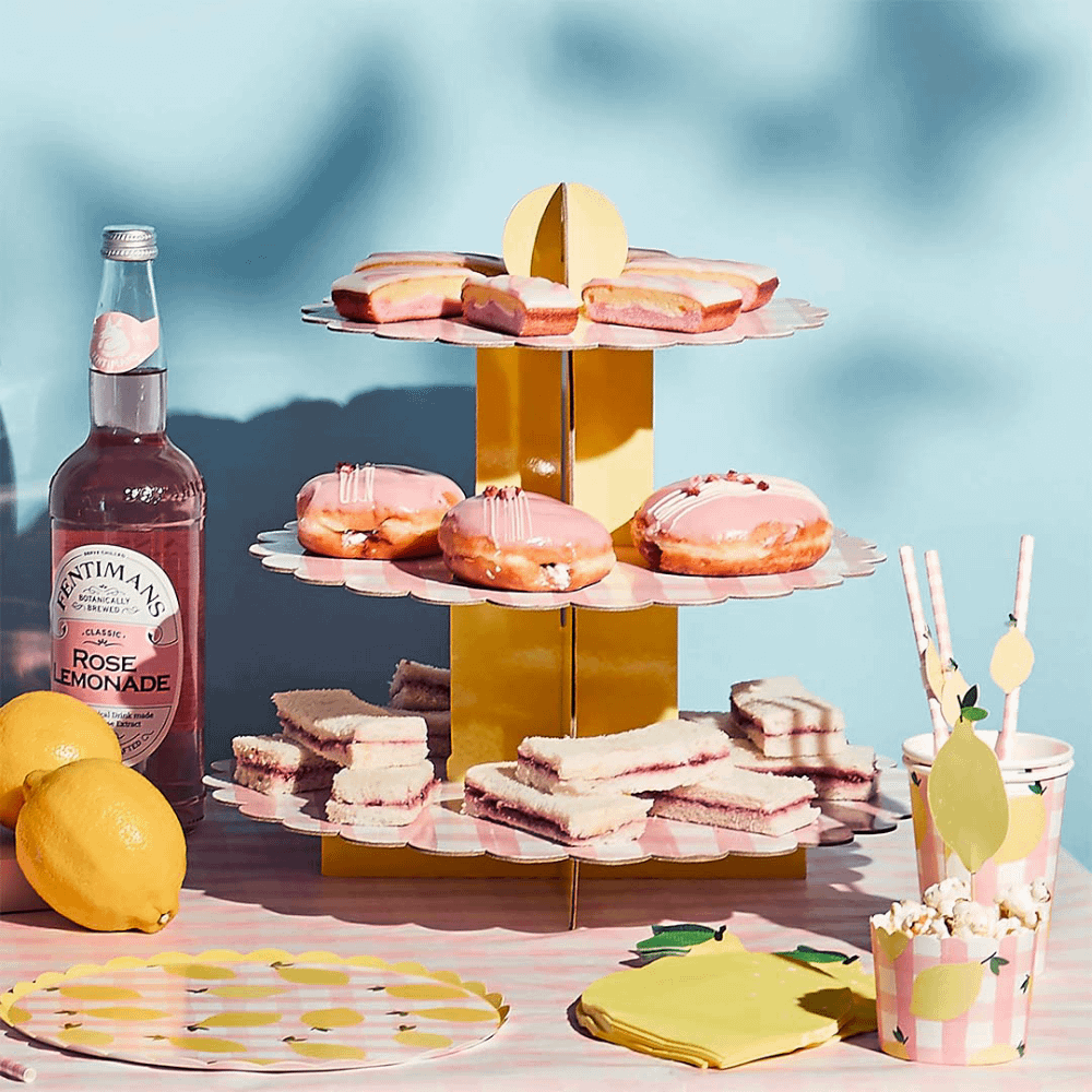 Cupcake plateau gevuld met snacks staat naast een roze flesje limonade en op een roze met wit geblokt tafelkleed