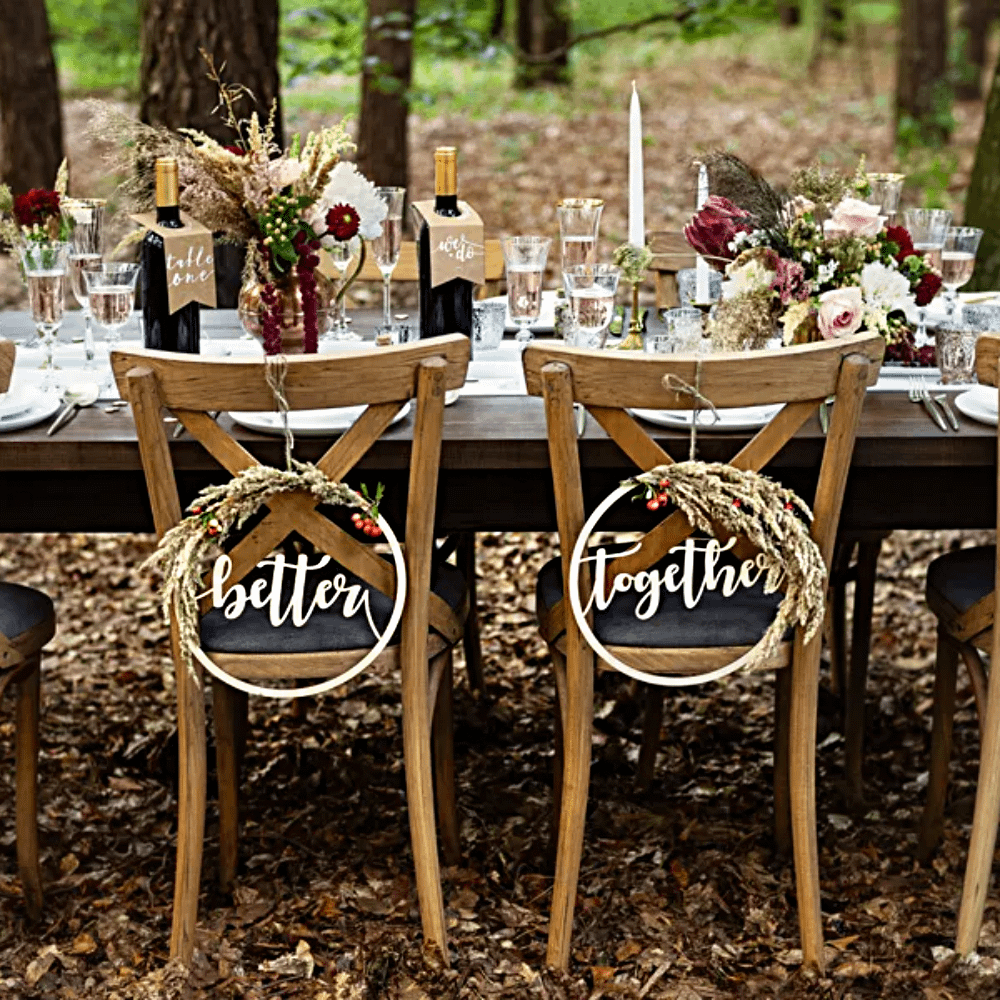 Houten tafel staat in een bos in de herft en is versierd met bloemen, een witte tafelloper en stoelhangers met de tekst better together