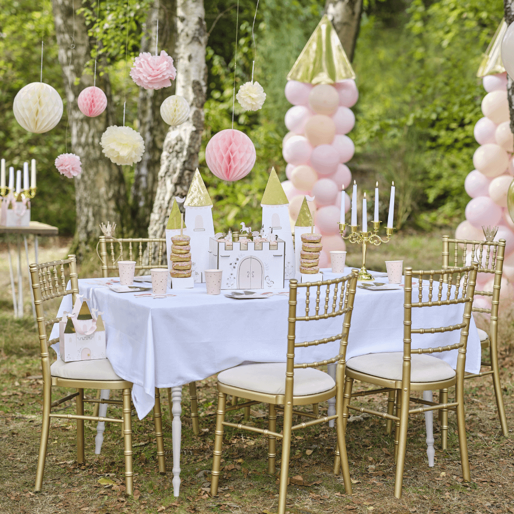 Tafel met wit tafelkleed en gouden stoelen staat in een bos omringd door prinsessenversiering bestaande uit donuts, roze ballonnen en honeycombs