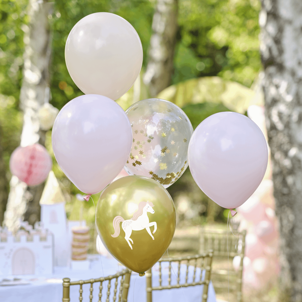 Lichtroze en perzikkleurige ballonnen zweven onder een boom voor een witte feesttafel en gouden stoelen