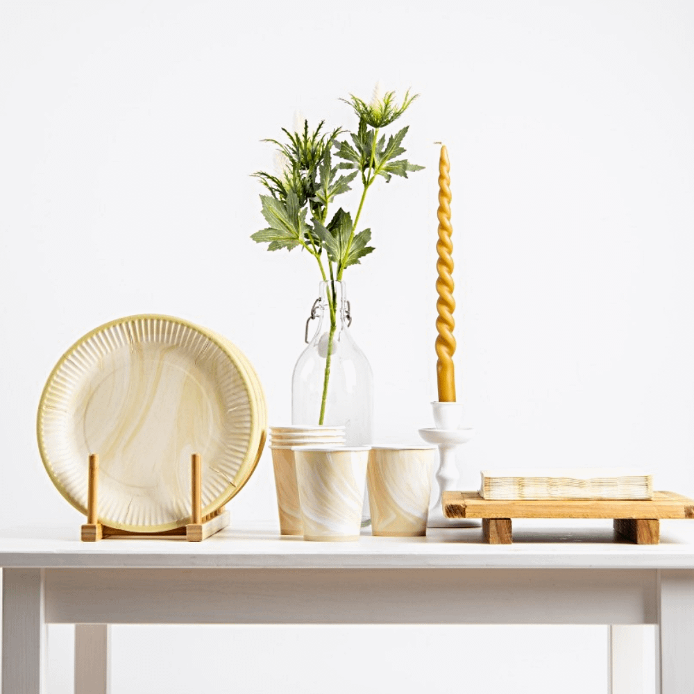 houten tafel versierd met marmeren beige en witte borden, bekers en servetten naast een gouden kaars en een glazen vaas met groen blad