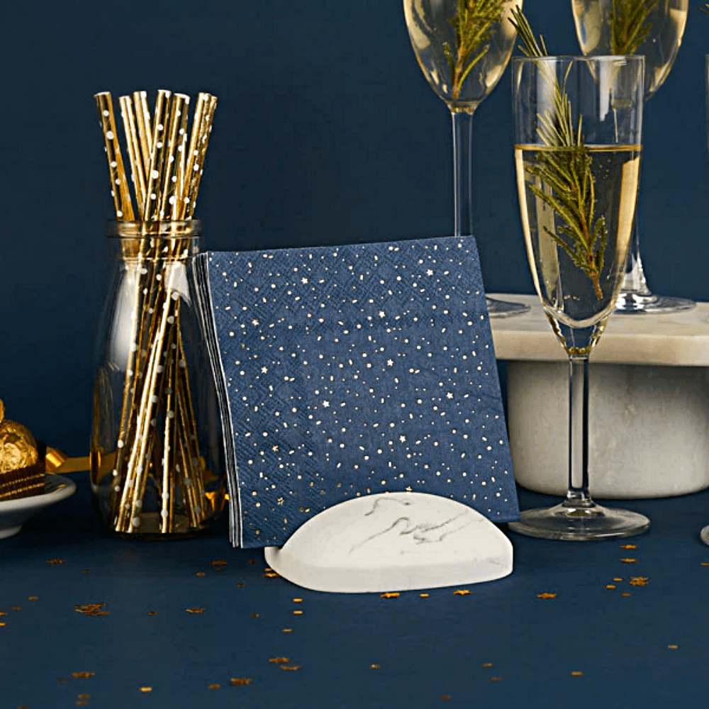 Donkerblauwe servettenmet gouden sterren, stippen en streepjes staan in een marmeren houder naast een champagneglas en gouden rietjes