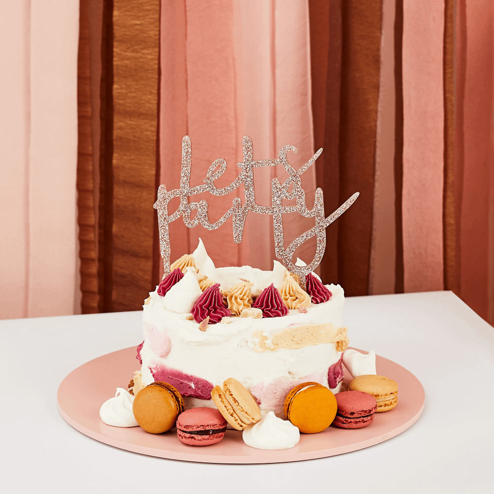 taart topper in het rose goud met glitters en de tekst lets party zit in een witte taart en is versierd met macarons