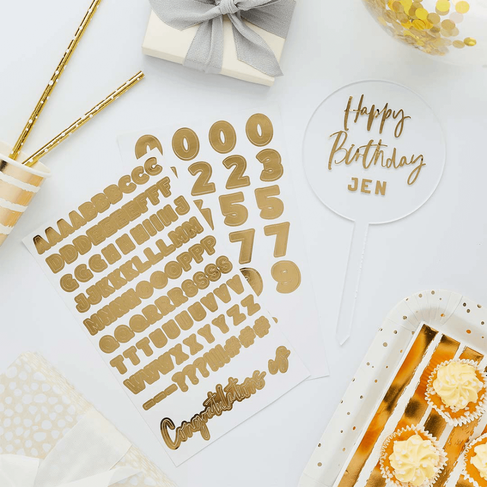gouden stickervellen met een acryl en transparant bordje liggen op een witte tafel naast cupcakes