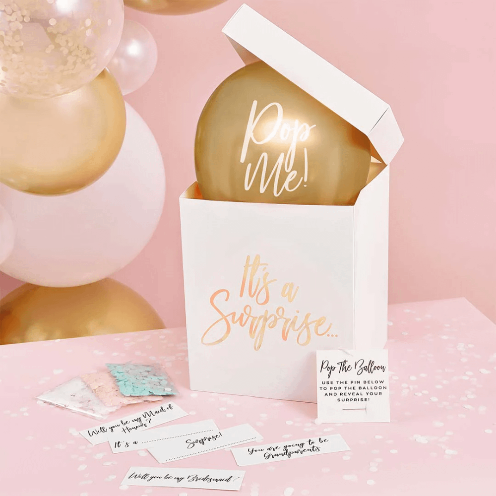 Witte doos met gouden tekst en metallic gouden ballon staat voor een roze muur op een witte tafel met blauwe en roze confetti