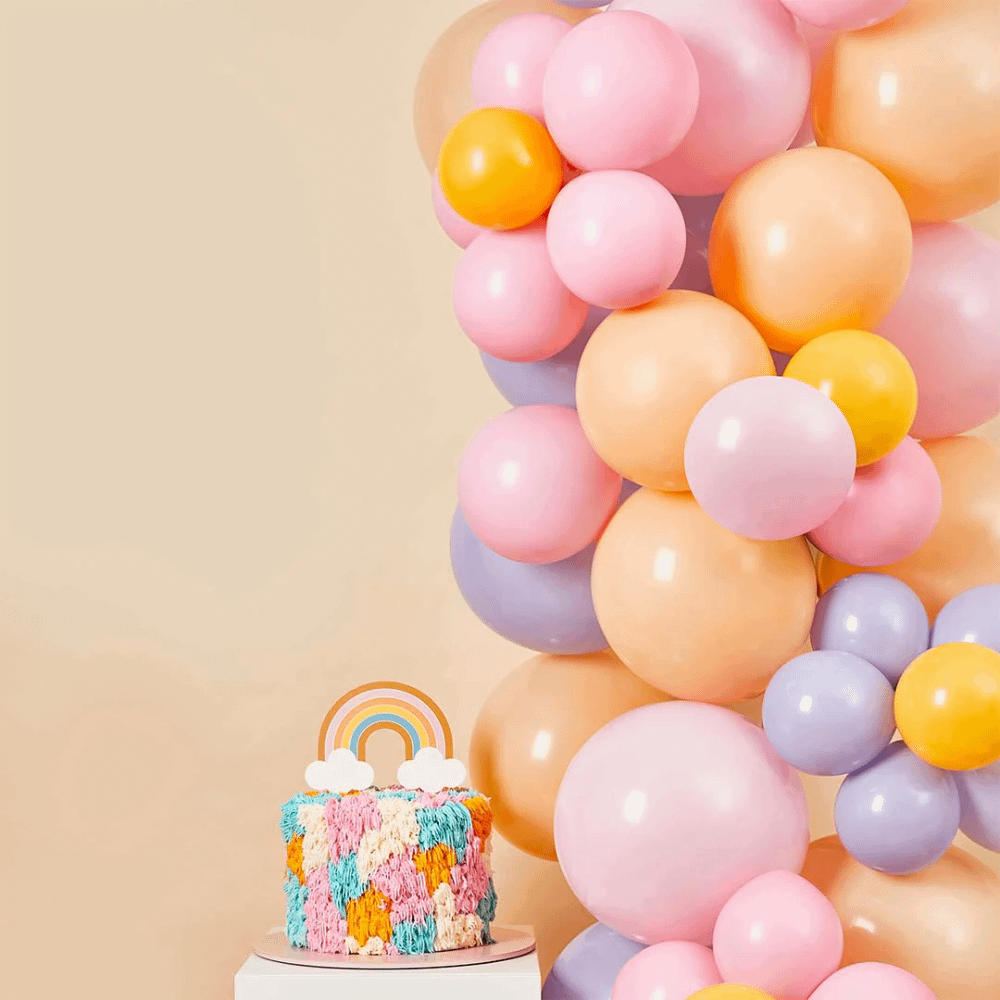 Ballonnenboog in pasteltinten en met bloemen staat voor een perizkkleurige muur naast een regenboog taart