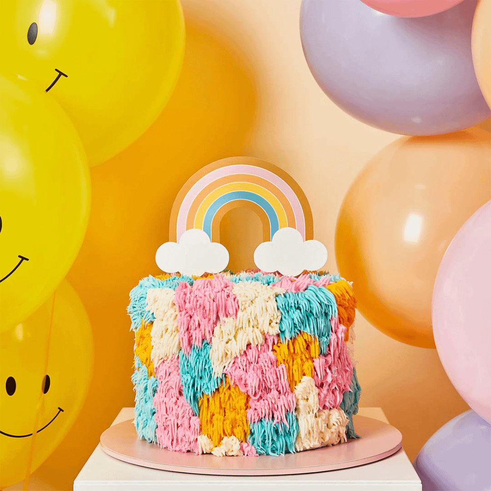 Regenboogtaart met taart topper staat voor een perzikkleurige muur met gele smiley ballonnen