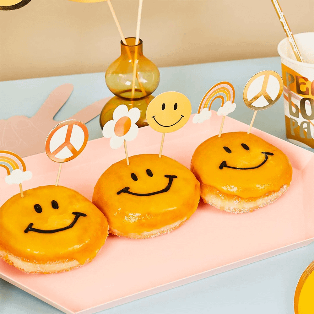 Donuts met gele smileys erop zijn versierd met sateprikkers met regenboogjes, madeliefjes en smileys erop en liggen op een lichtroze plank voor een glazen vaasje