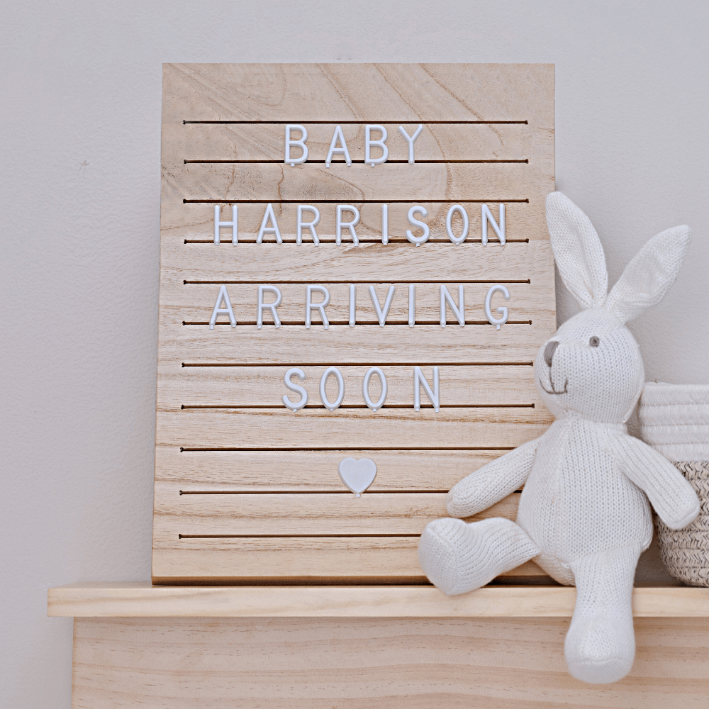 houten letterbord met witte letters staat op een plank in een babykamer naast een mandje en een wit knuffel konijn