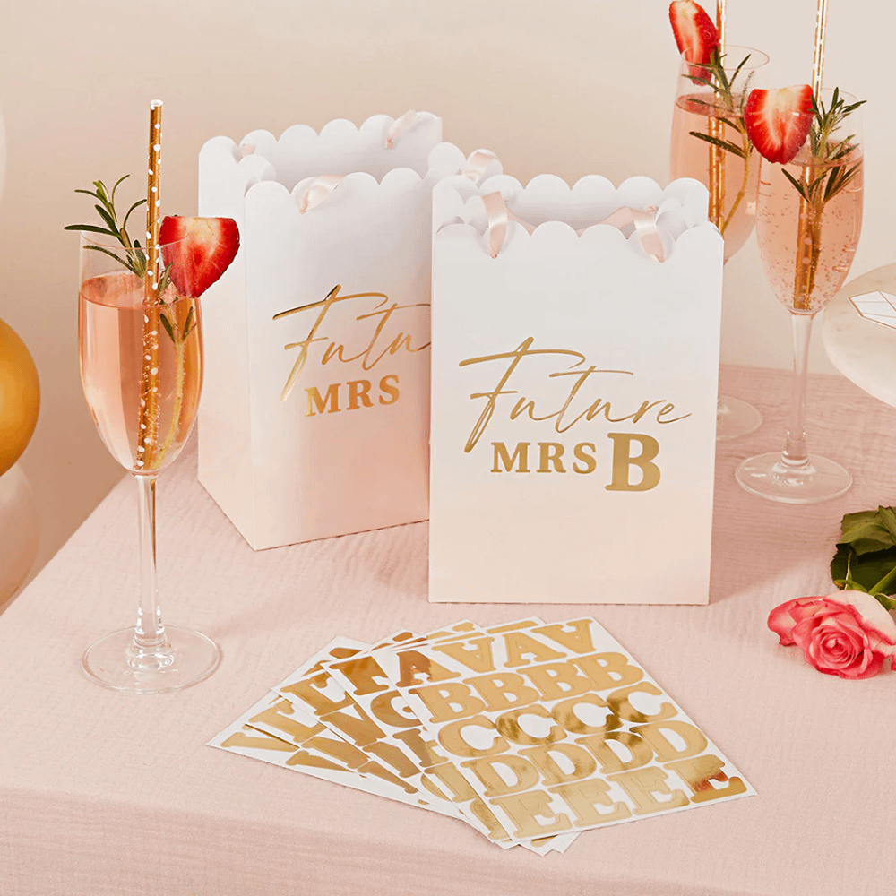 perzikkleurige en witte cadeautasjes met ombre effect en gouden tekst future mrs staan op tafel naast een glas champagne met een aardbei