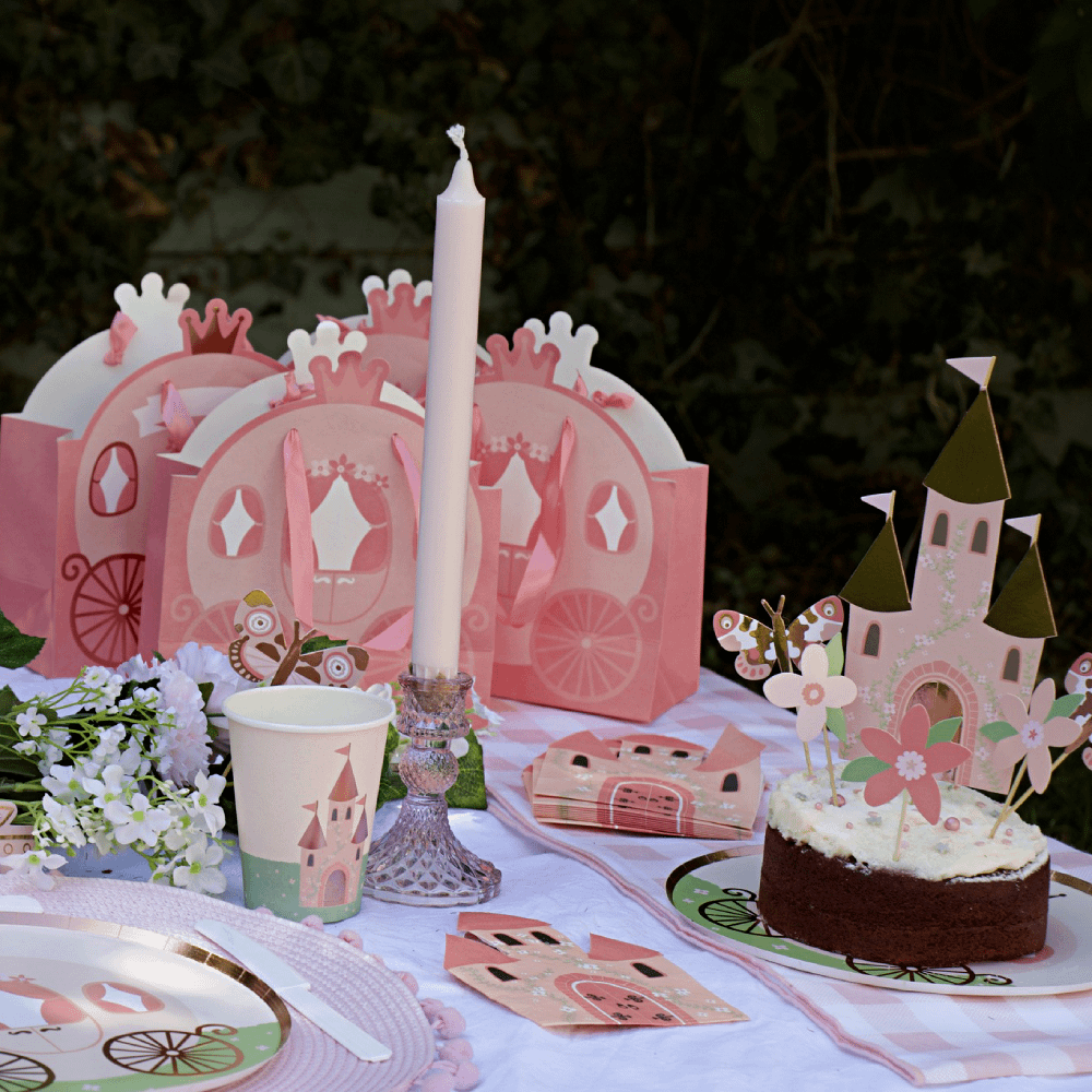 Tafel versierd met een roze tiara, bordjes met een koets erop en cadeautasjes in de vorm van een koets naast een bloemenslinger en een lange kaars