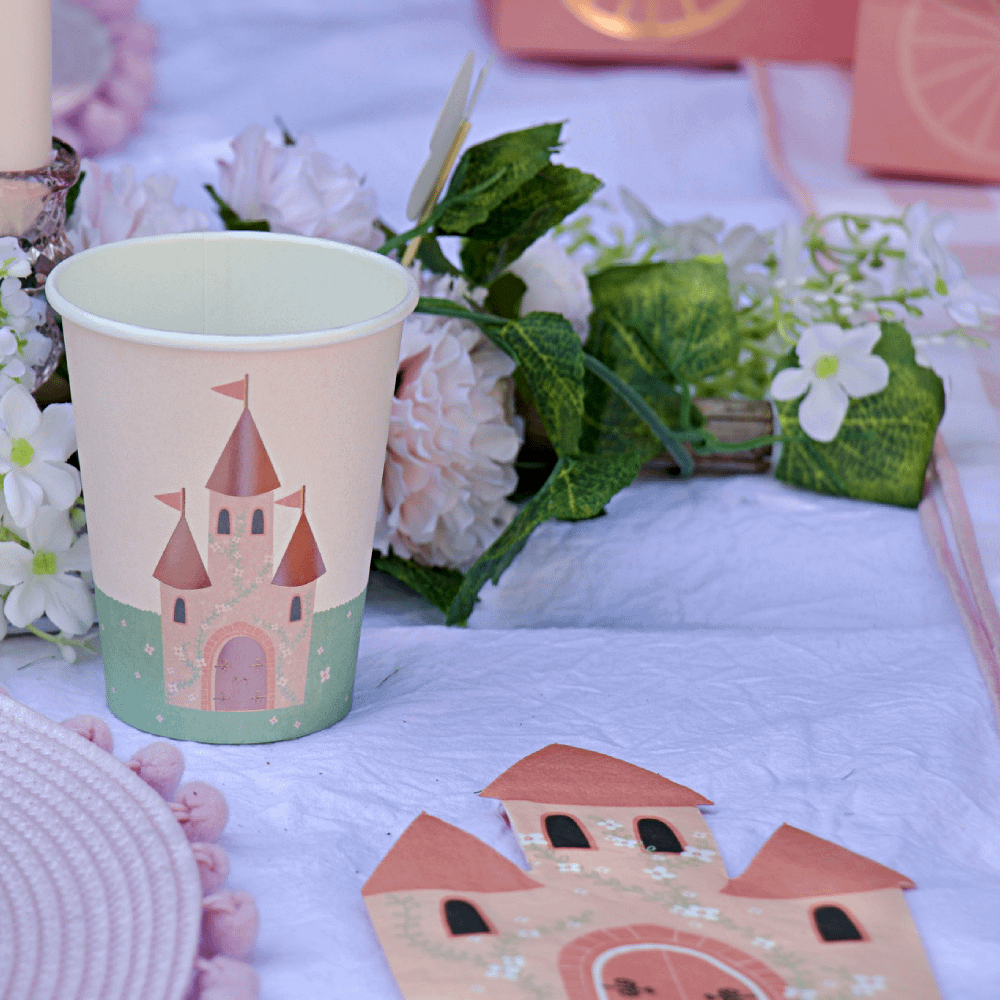 Papieren bekers en servetten in de vorm van een kasteel liggen op tafel naast een bloemenkrans met roze bloemen