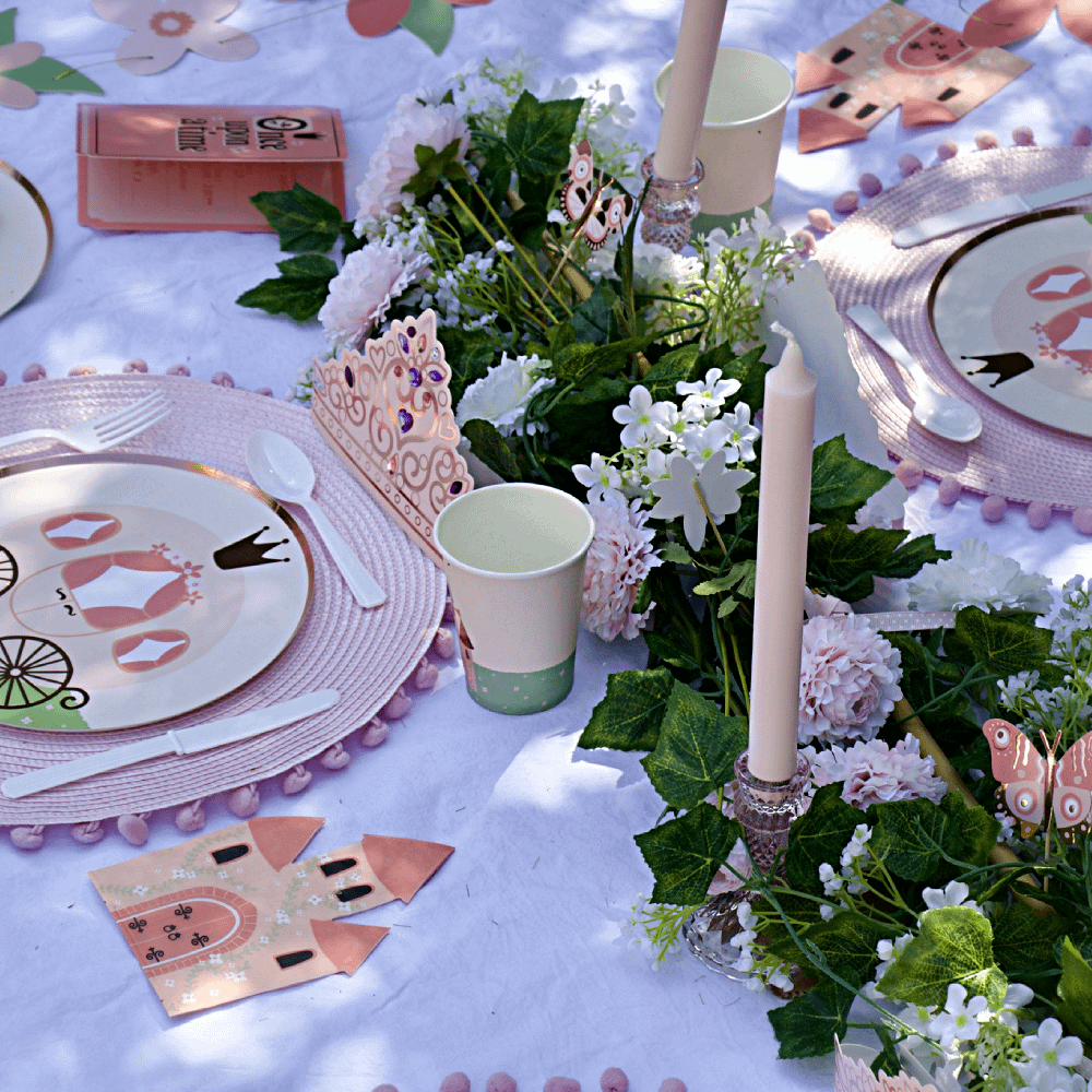 Tafel versierd met een roze tiara, bordjes met een koets erop en cadeautasjes in de vorm van een koets naast een bloemenslinger en een lange kaars