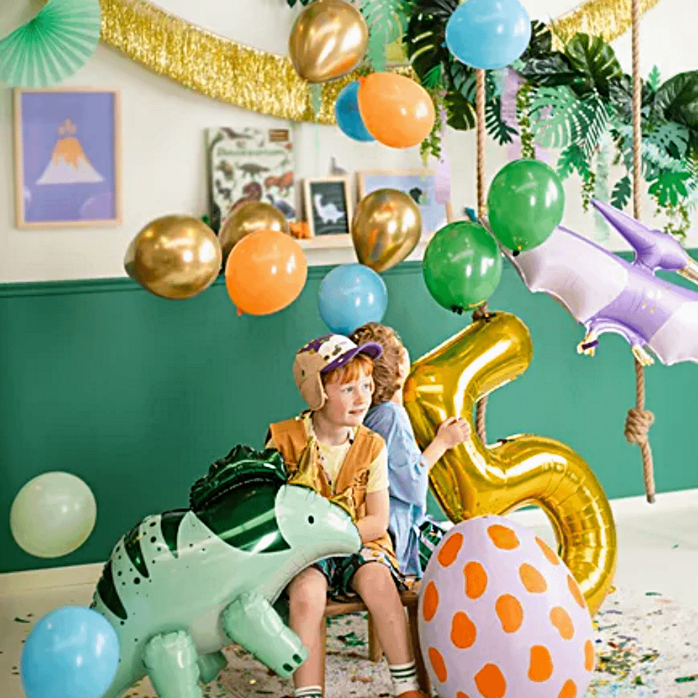 twee jongetjes met dino ballonnen en een gouden cijfer 5 zitten tussen oranje, blauwe en groene ballonnen onder een palmbladeren slinger