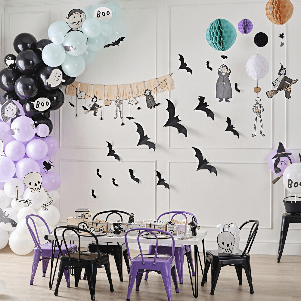Kamer versierd met een zwarte tafel en zwarte en paarse stoelen, papieren vleermuizen aan de muur en een zwart, paars en mintgroene ballonnenboog voor halloween