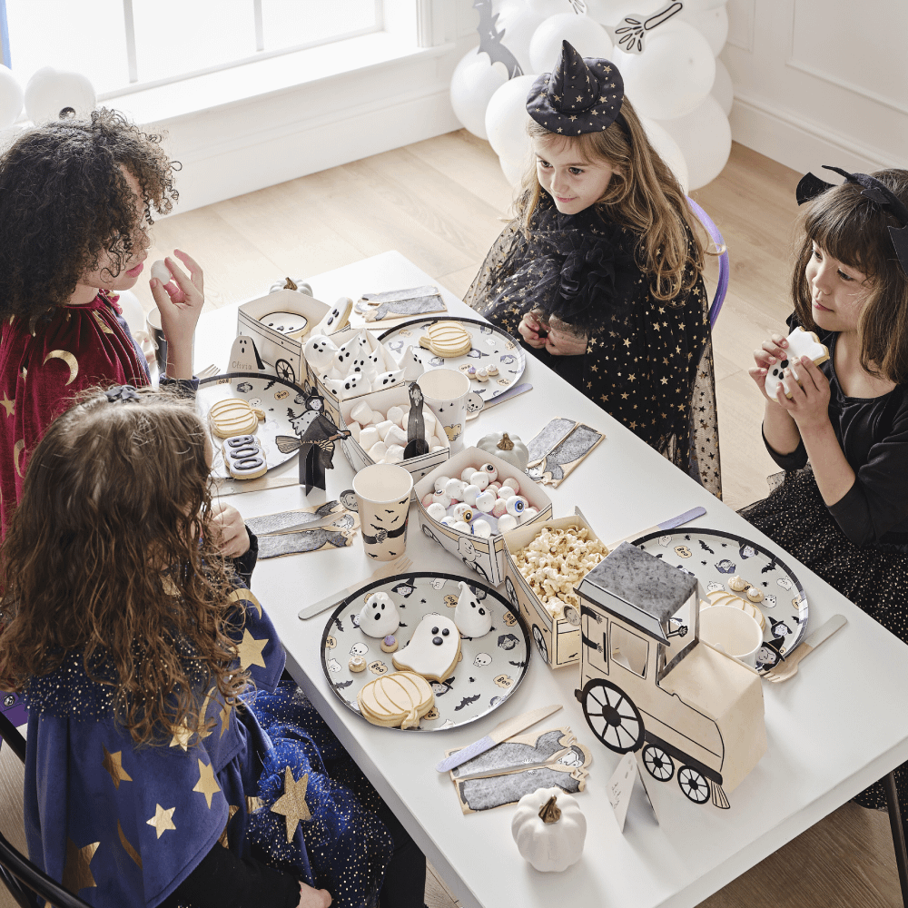 Kinderen in halloween kostuums zitten aan een tafel versierd met een treintje met snacks, bordjes en bekers met halloween poppetjes erop