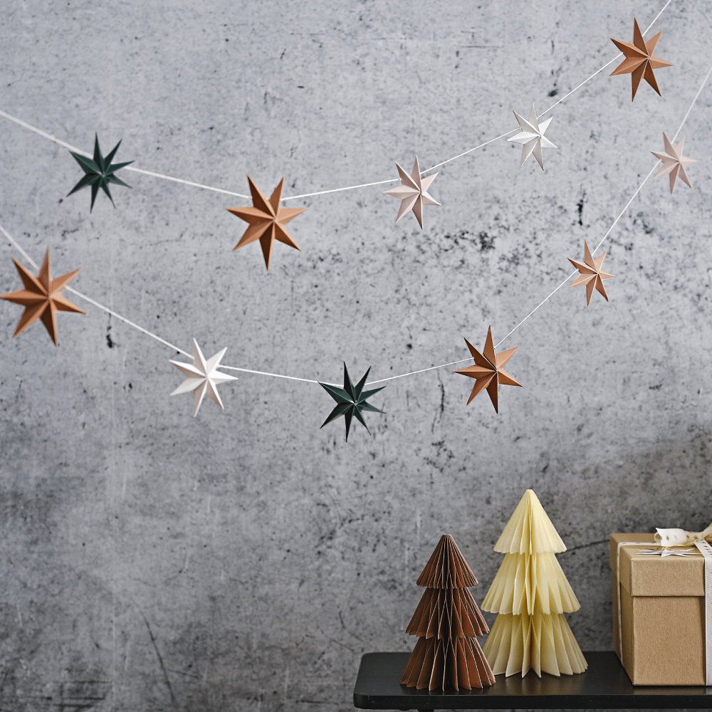 Slinger met 3D sterren in het koper, donkergroen, grijs, wit en beige hangt voor een grijze muur boven twee honeycomb kerstbomen