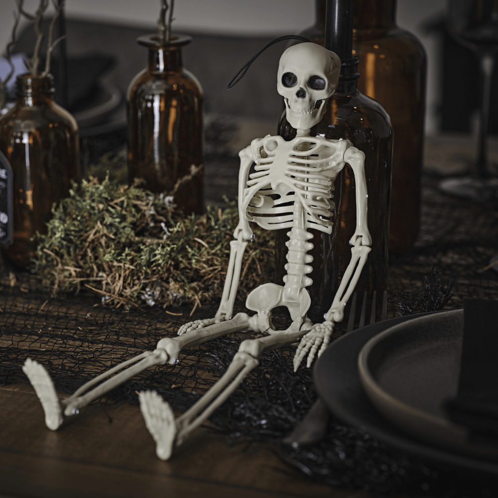 Wit, plastic skelet zit op een houten tafel op een zwarte tafelloper naast donkergroen mos