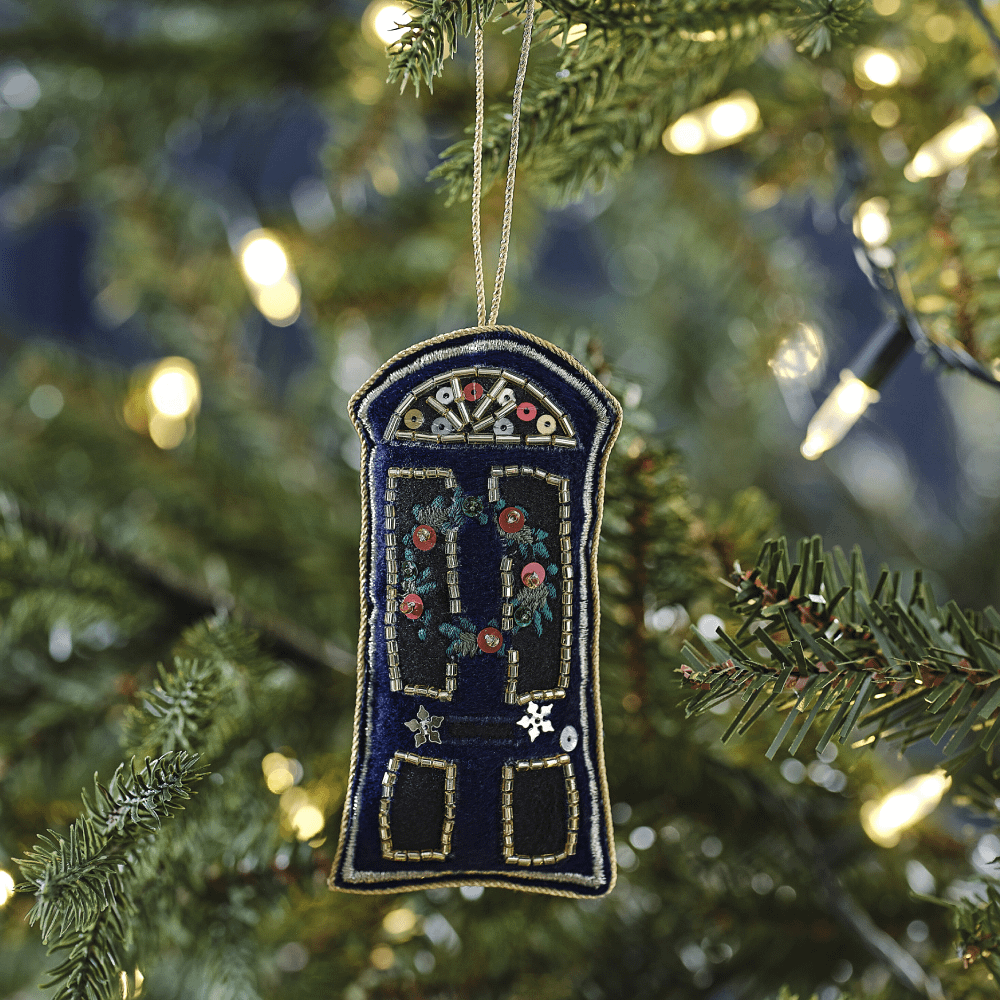 Kerstboomhanger in de vorm van een marineblauwe deur met kerstkrans en gekleurde kralen hangt in de kerstboom met lampjes