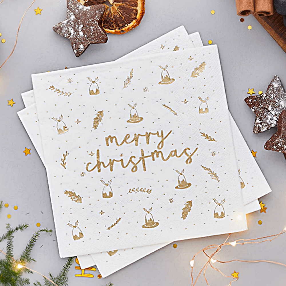 Witte servetten met gouden print en de tekst merry christmas liggen naast een lampjesslinger en een sinaasappelschijfje