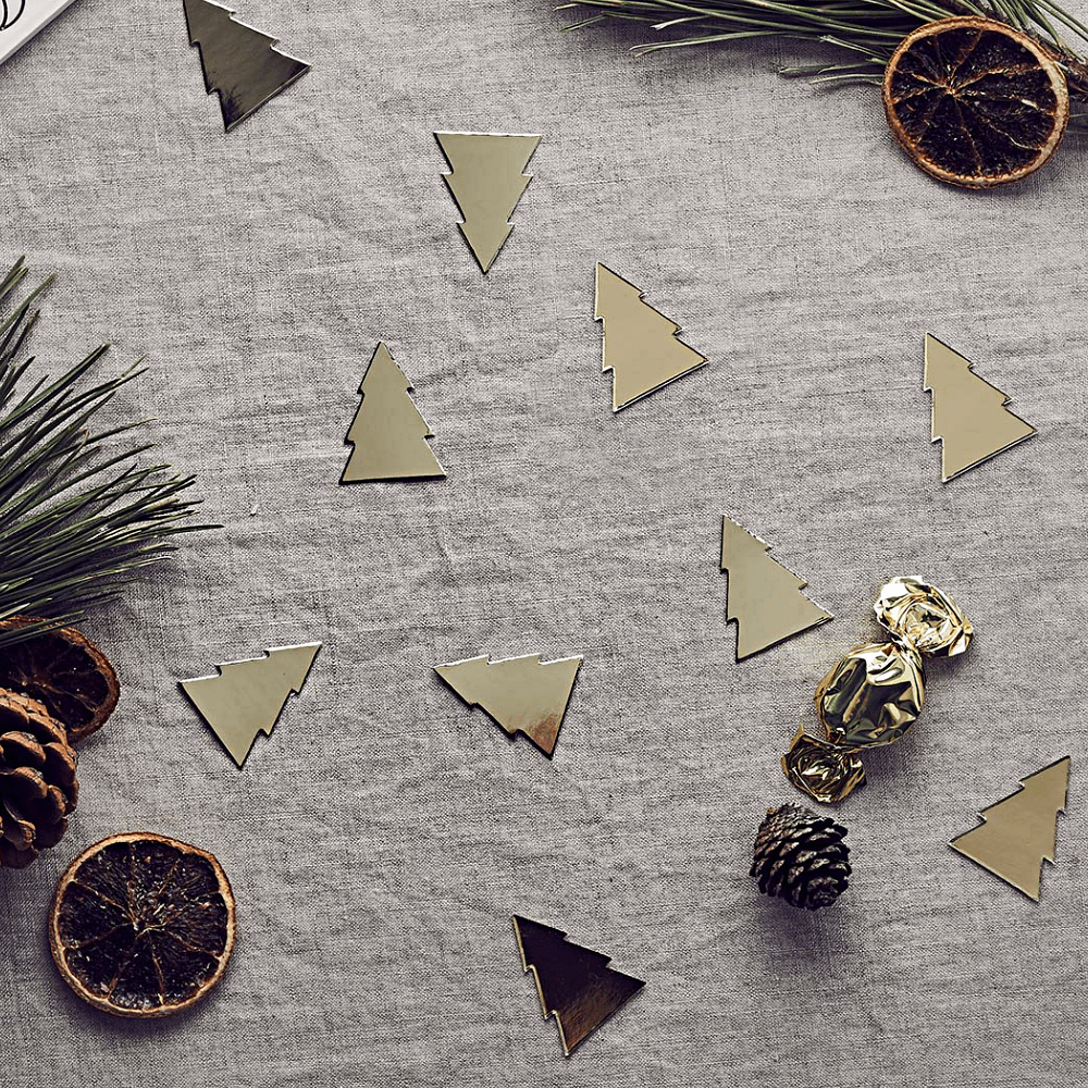 Confetti in de vorm van gouden kerstboompjes liggen op een katoenen kleed versierd met gouden bonbons, dennenappels en dennentakken