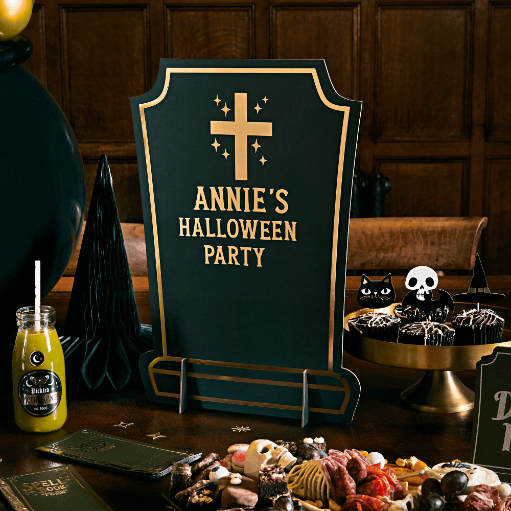 Bord in de vorm van een grafsteen met gouden tekst staat op een tafel vol halloween versiering
