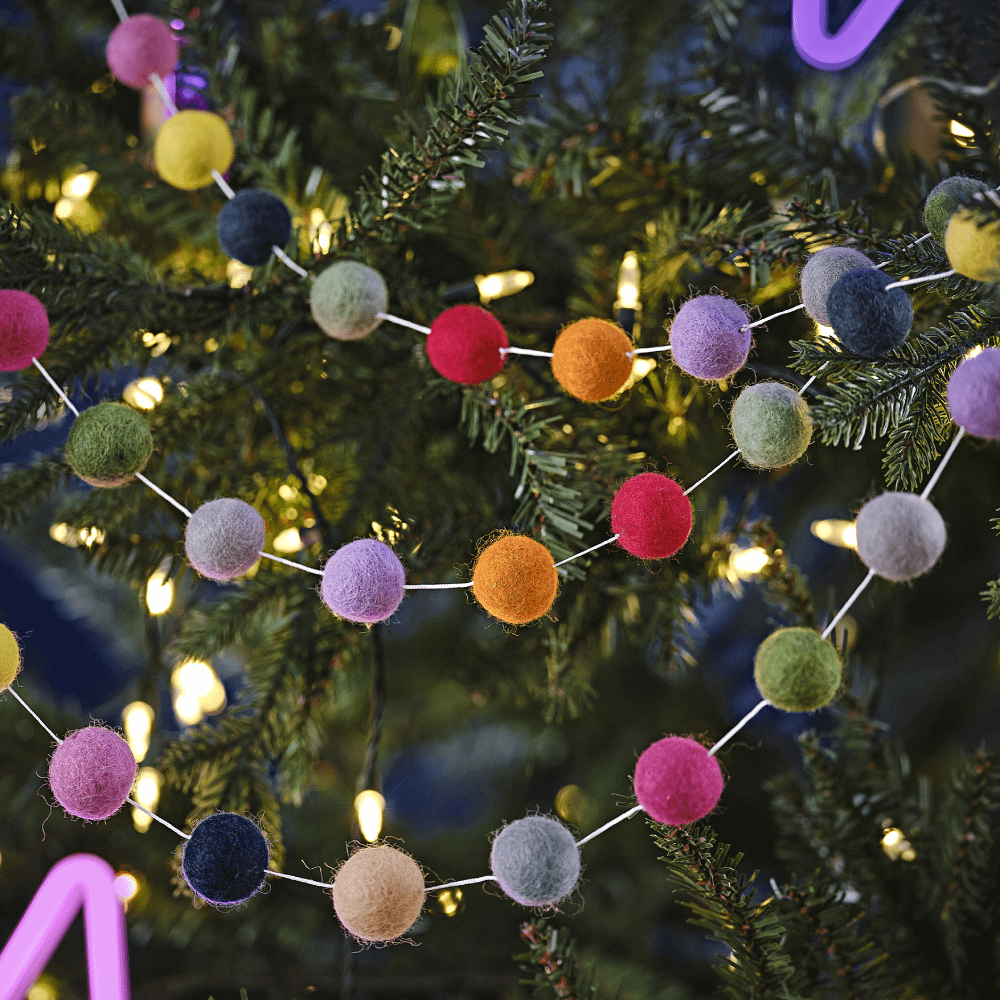 Slinger van vilt met pom poms in de kleuren saliegroen, roze, beige, marinablauw, perzik en grijs hangt in de kerstboom