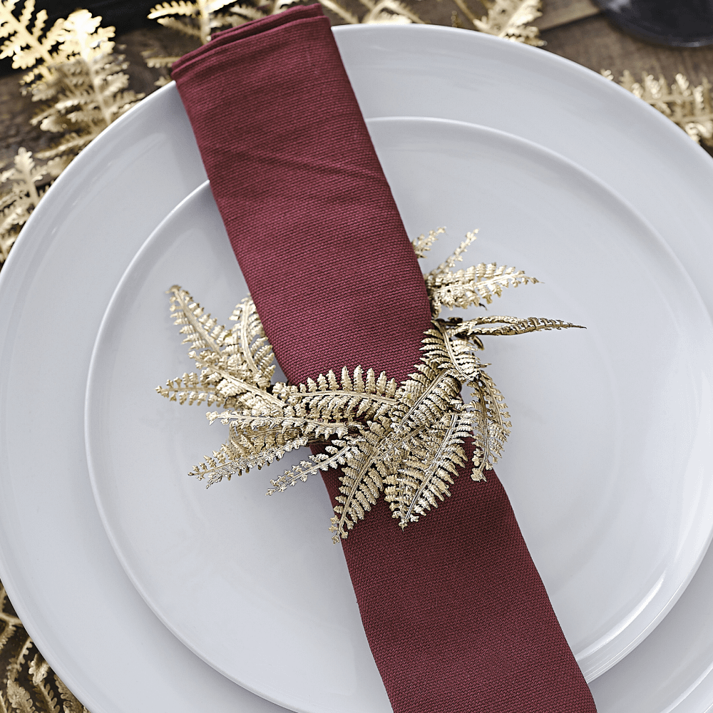 Servetring met bruine binnenkant en gouden bladeren van een varen zit om een rode servet heen en ligt op een wit bord