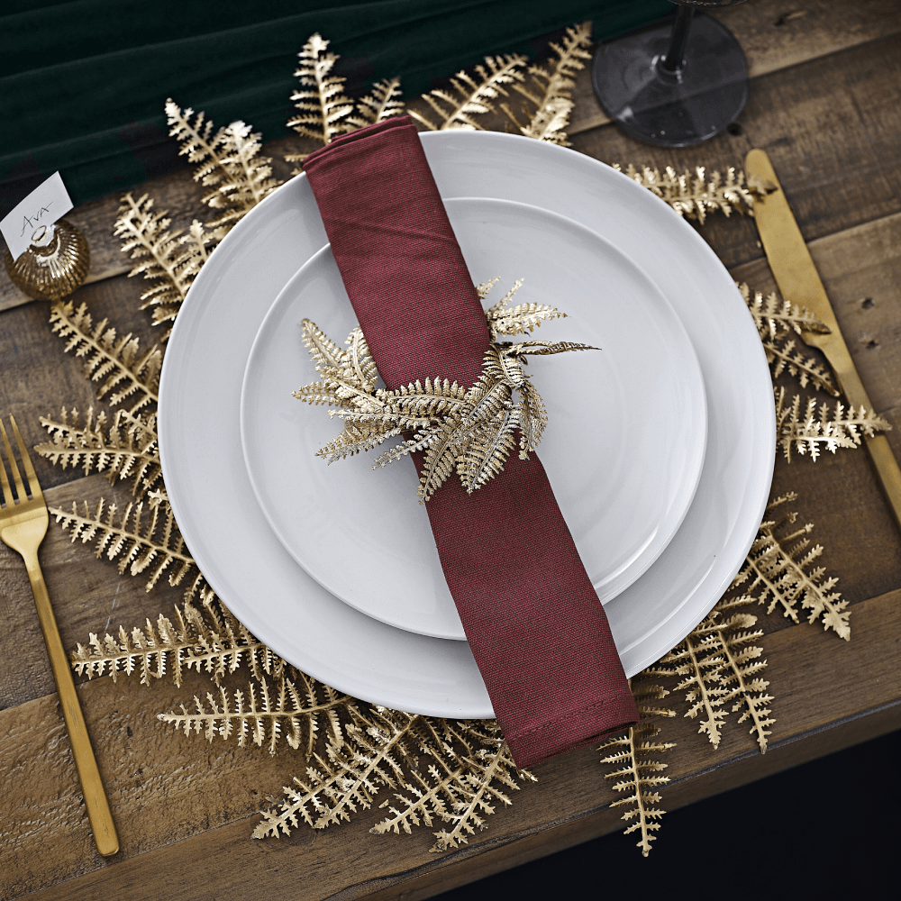 Placemat met gouden varen ligt op een donkerbruine tafel onder een wit bord met rode servet en matchende servetring
