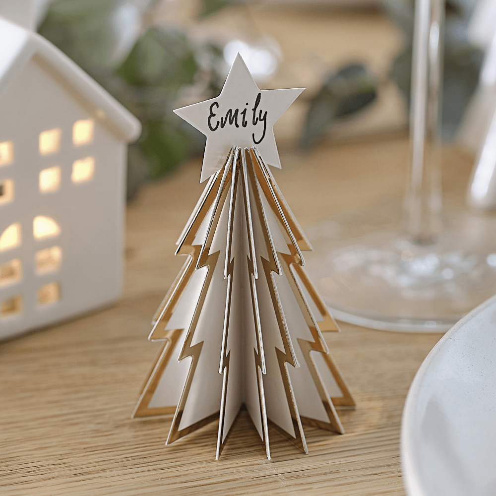 Witte kerstboom van karton met gouden rand en een ster als piek staat op een houten tafel voor een wit huisje