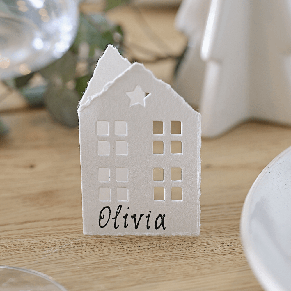 Wit tafelkaartje in de vorm van een huisje met ramen en een ster staat op een houten tafel en is beschreven met de naam olivia