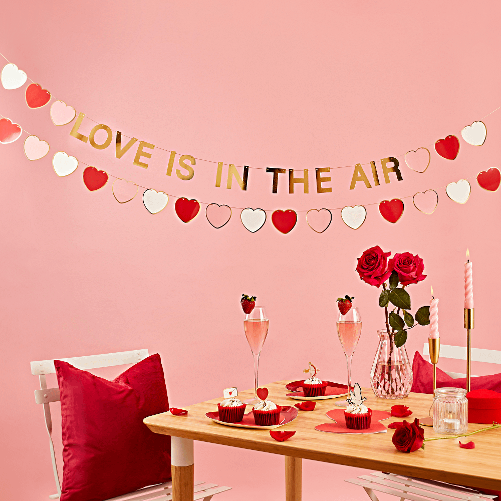 Gouden slinger met de tekst love is in the air en hartjes in het wit, roze en rood hangt boven een houten diner tafel