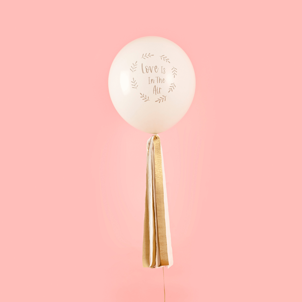 Ballon met gouden tassels en de tekst love is in the air hangt voor een roze muur