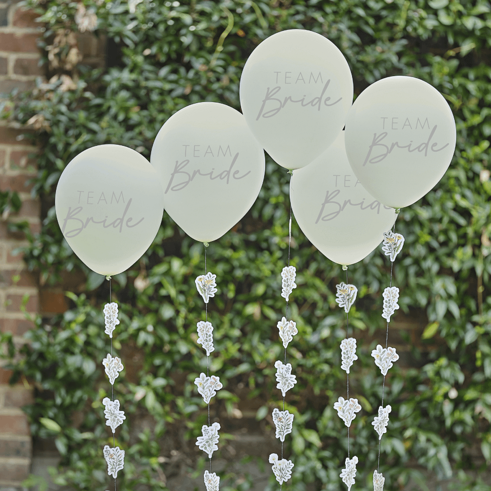 Saliegroene ballonnen met grijze tekst team bride en bloemen en planten hangen voor een heg met groene bladeren
