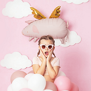 Folieballon in de vorm van een roze varkentje met gouden vleugels zweeft achter een meisje met een roze zonnebril voor papieren wolkjes