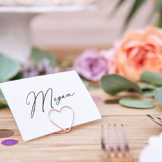 Rosé gouden hartvormige tafelkaart houder met wit kaartje en bloemen op de achtergrond