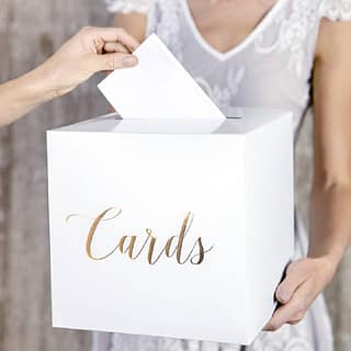 Vrouw houdt witte kaartendoos vast met gouden letters erop en iemand doet er een envelop in