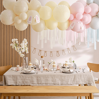 Houten tafel met ivoorkleurig tafelkleed en paasversiering in verschillende pasteltinten bestaande uit ballonnen, eieren en een houten slinger