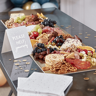 Gouden hapjesplank gevuld met olijven, pretzels, druiven en dadels ligt op een reflecterende tafel omringd door gouden confetti