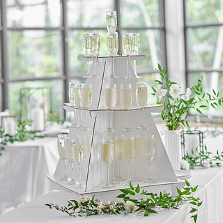 Witte drankjesstandaard met drie verdiepingen gevuld met champagneglazen staat op een witte tafel met witte bloemen en groene takken met bladeren