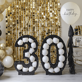 Ballonstandaard cijfers 30 in het zwart met witte ballonnen staat voor een gouden backdrop en naast een reuzeballon met zwarte franjes