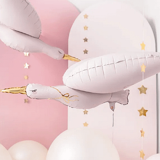 Folieballon in de vorm van een ooievaar met lichtroze gloed en gouden details zweeft in een roze babykamer met gouden sterretjes